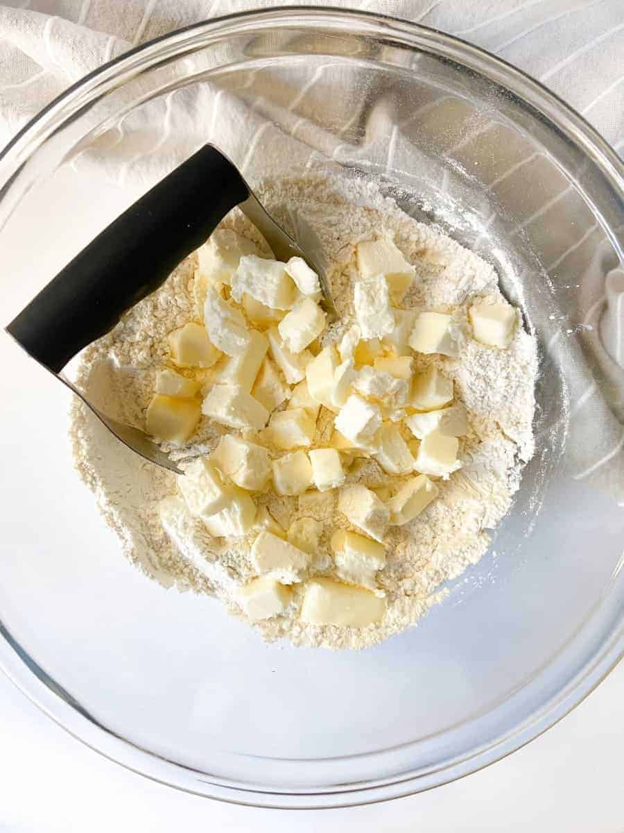A large bowl filled with dry ingredients, cubed butter, and a pastry cutter.