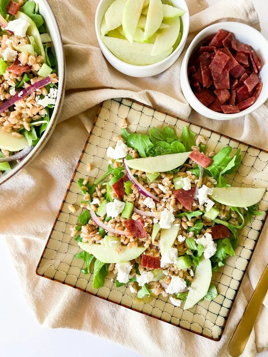 A plate of apple farro and bacon salad with two small bowls filled with extra ingredients. 