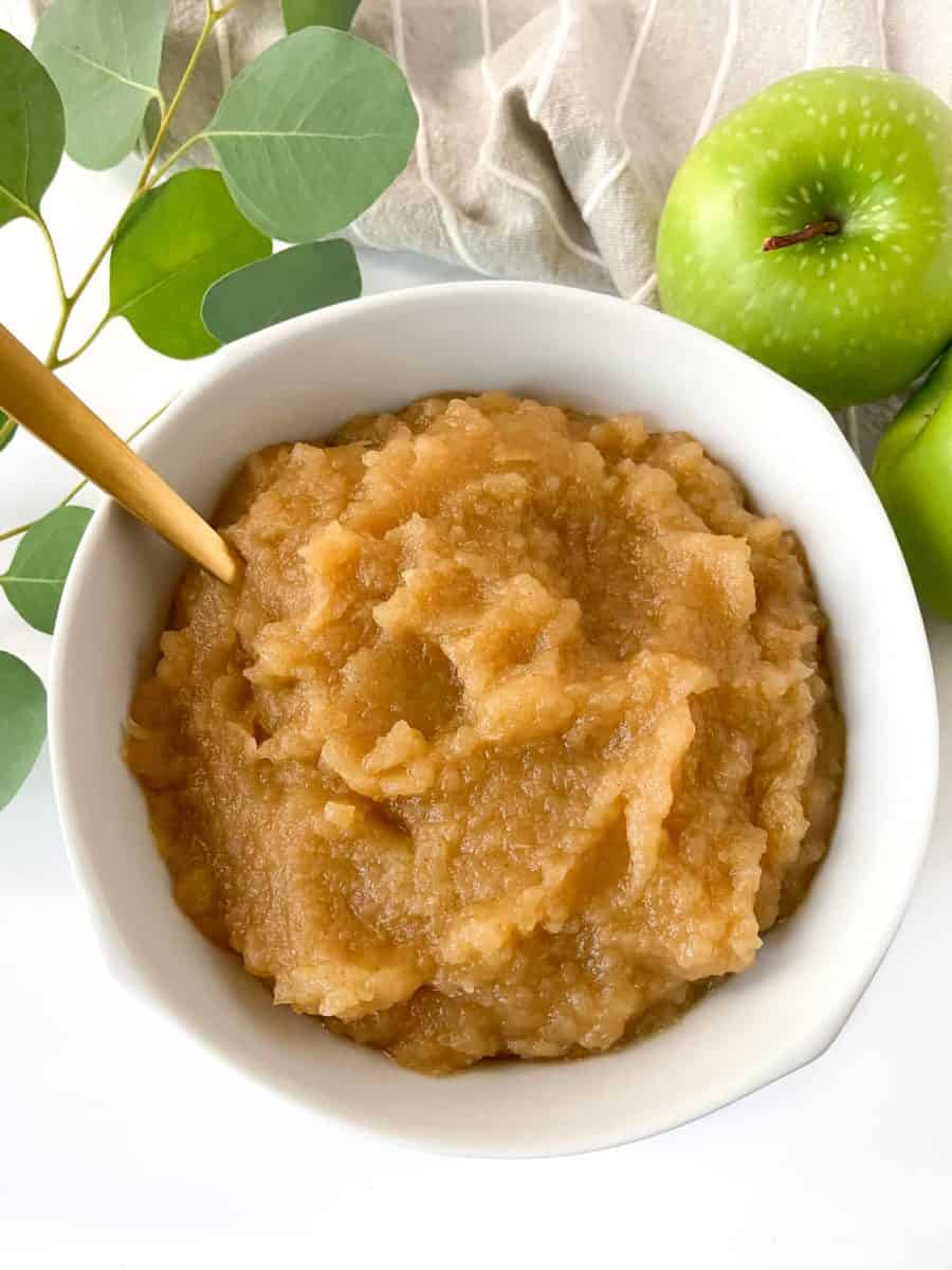 A white bowl of completed apple sauce with a golden spoon and green apples in the background. 