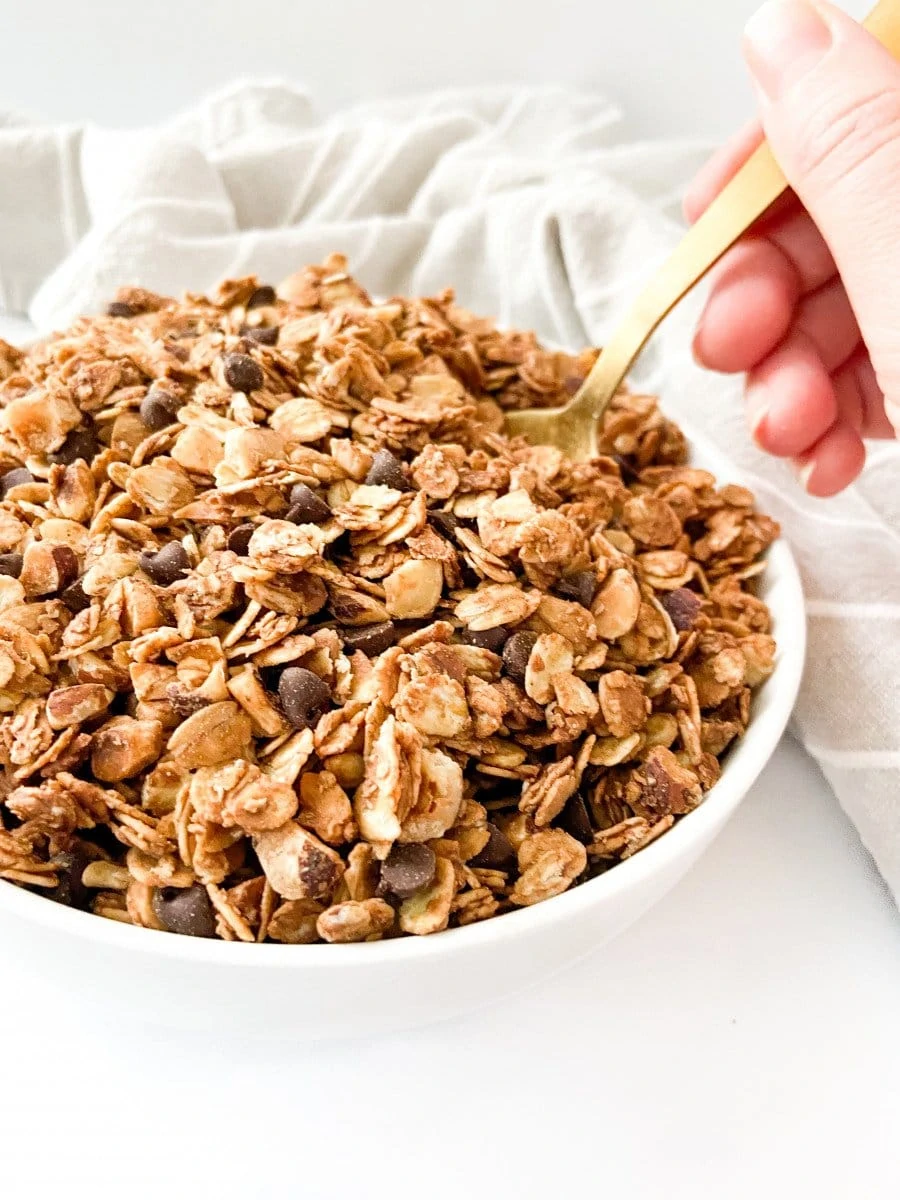A bowl of chocolate hazelnut granola with a hand scooping into it with a golden spoon. 