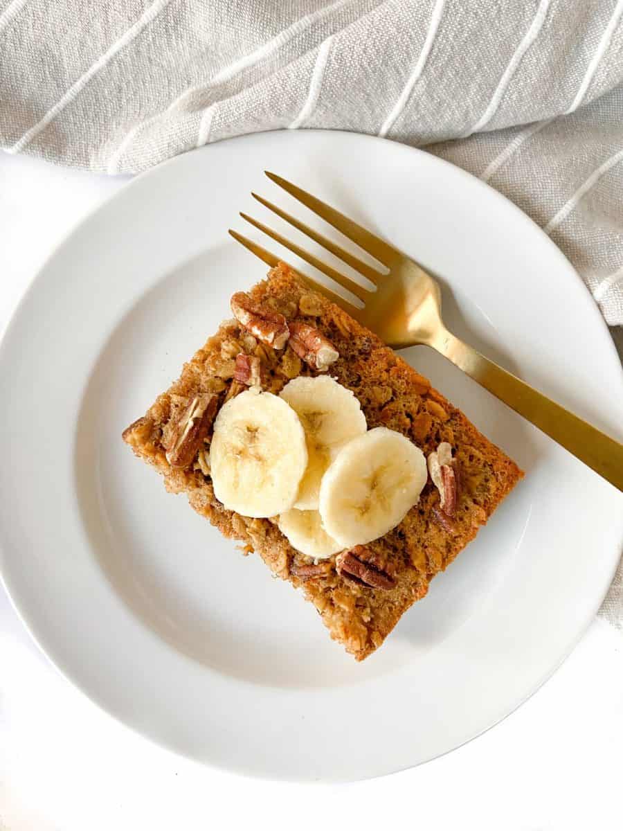 A single slice of baked oatmeal topped with banana slices, alongside a gold fork. 