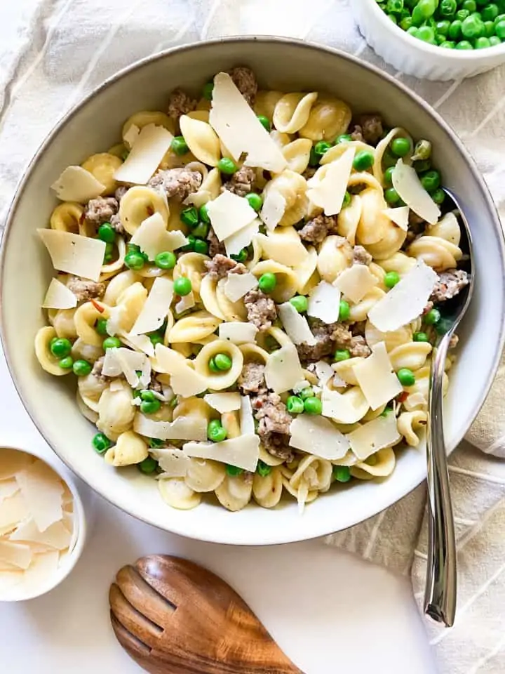 Orecchiette with Sausage and Peas in a bowl garnished with shaved Parmesan cheese, surrounded by a small bowl of extra cheese and peas.