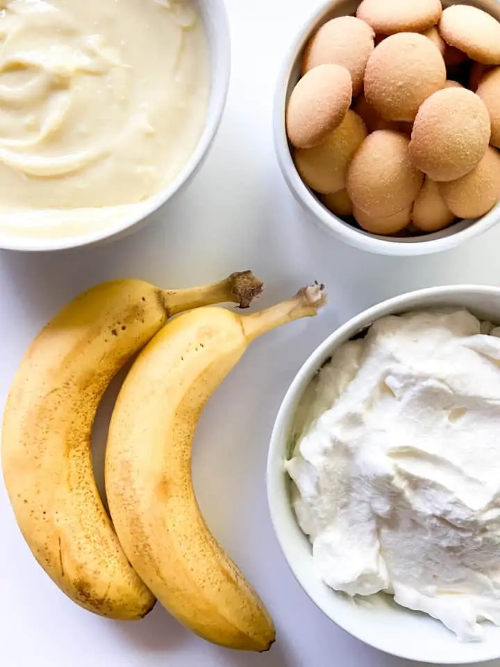 A bowl of pastry cream, a bowl of whipped cream, a bowl of mini vanilla wafer cookies, and two ripe bananas. 