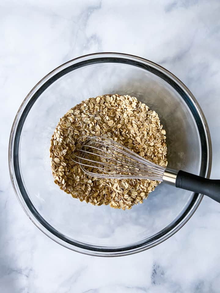 A clear bowl with oats, sugar, baking powder, and spices with a whisk.