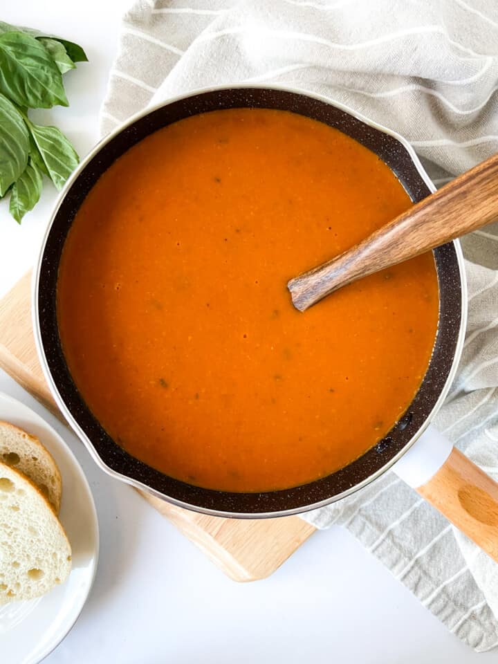 Tomato soup in a small saucepan surrounded by a kitchen towel, fresh basil, and bread.