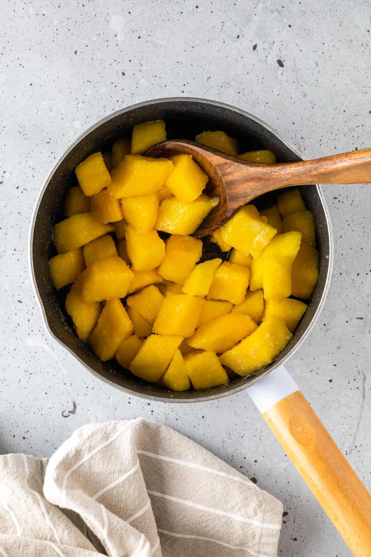 Sugar and lime juice-coated mango chunks in a small saucepan with a wooden spoon.