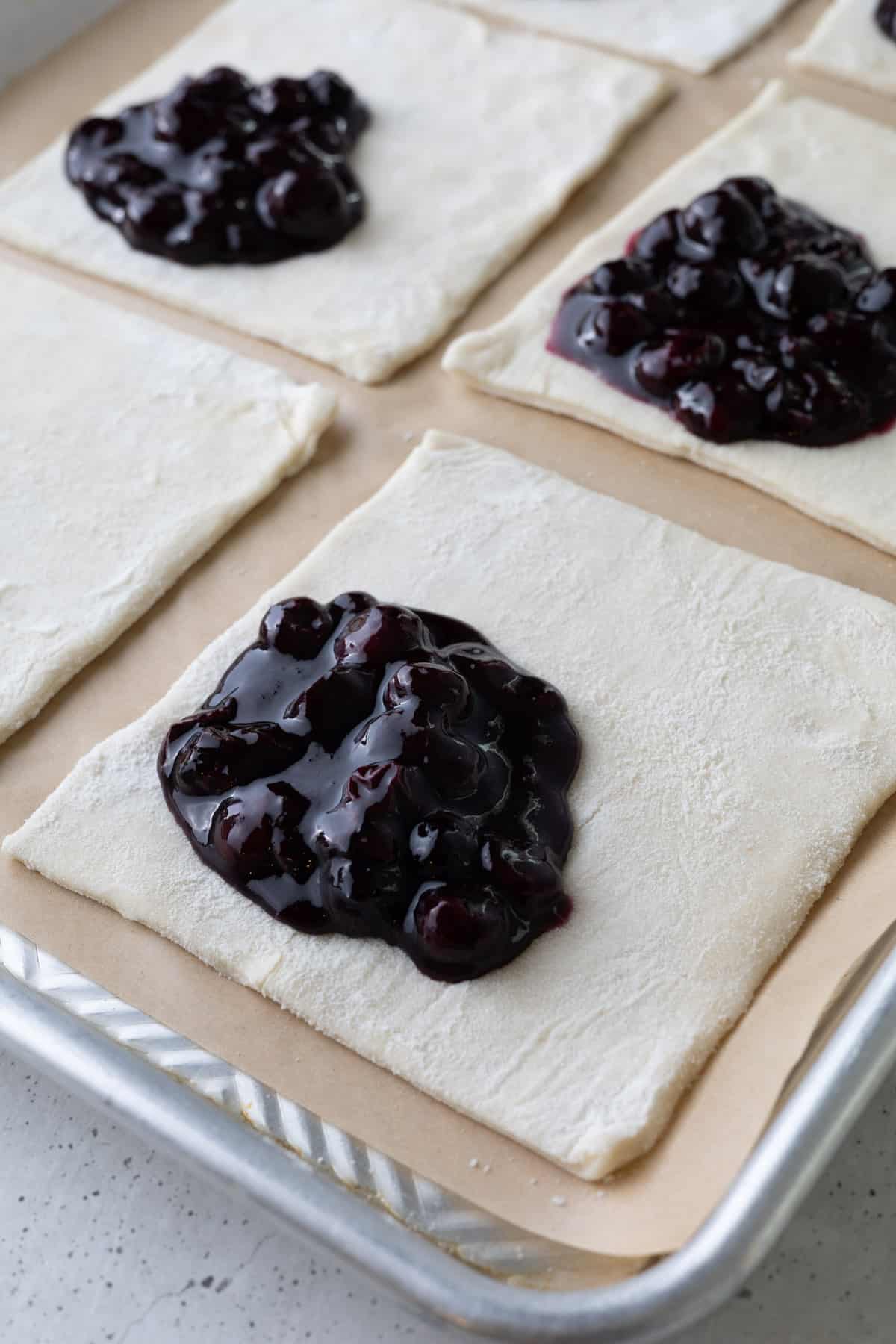 Blueberry filling-topped puff pastry squares on a parchment-lined baking tray.